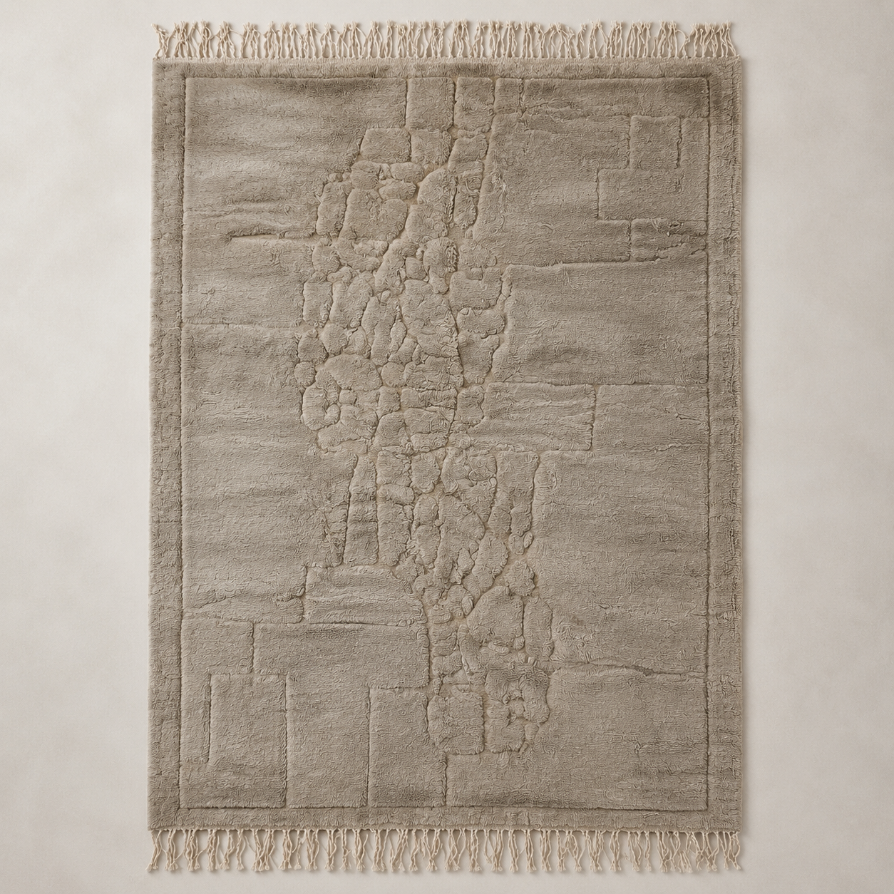 A rectangular area rug with a textured, abstract stone-like pattern in warm greige and beige tones, featuring fringed edges on all sides, shown flat against a neutral background.