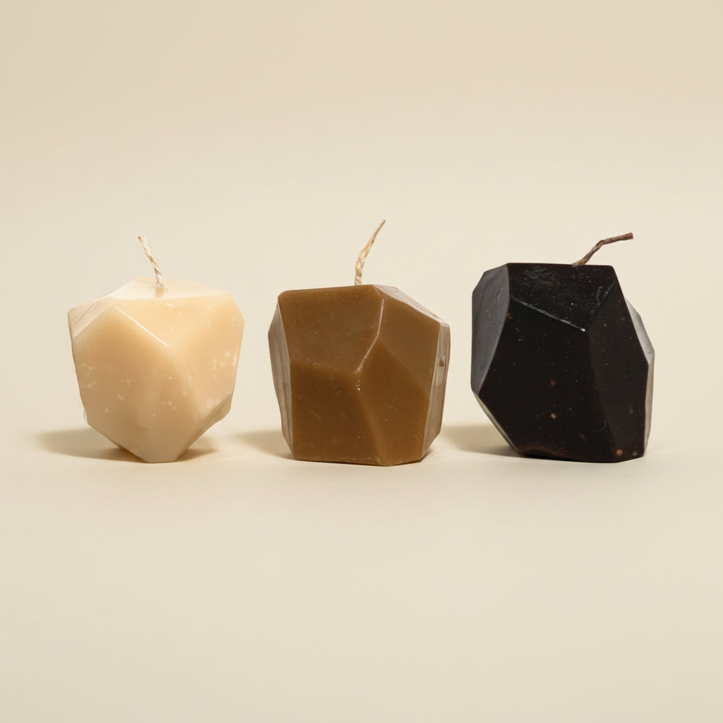 Three faceted candles in beige, brown, and black on a textured white background