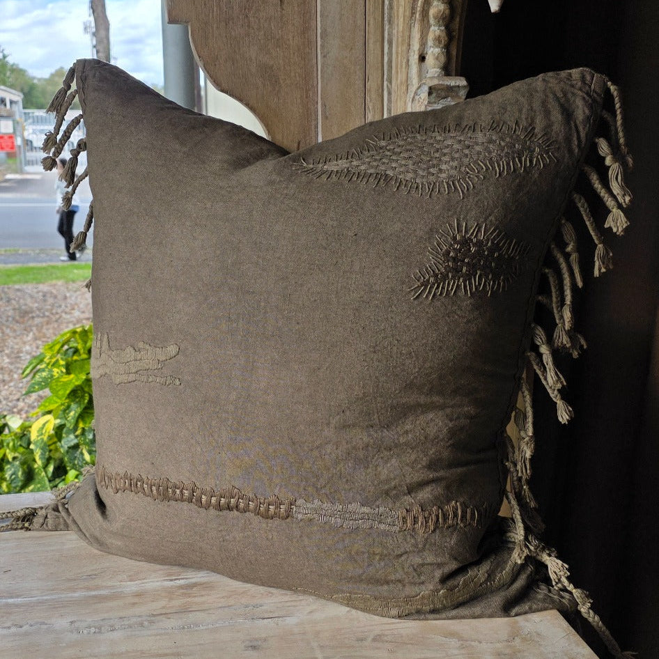 A square olive brown woven throw cushion with braided fringed tassels, tonal textured embroidered detailing in abstract stitching for an understated luxurious and elevated feel.