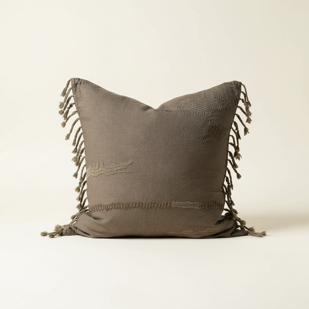 A square throw pillow in a textured olive taupe fabric with embroidered details and fringed tassels on the sides, set against a plain off-white background.