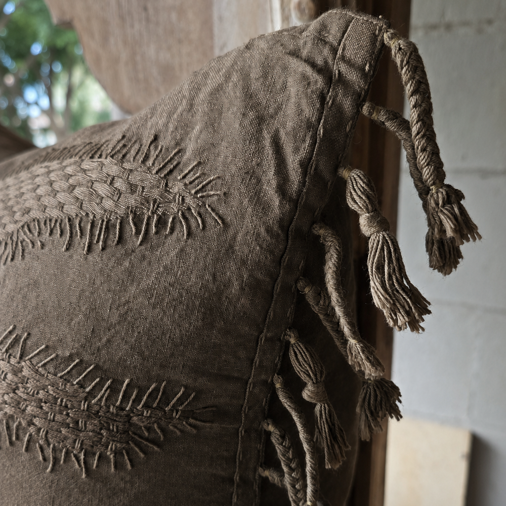 A close-up of a textured taupe throw pillow with woven stitching embroidery and hanging tassels, leaning against a rustic wooden frame.