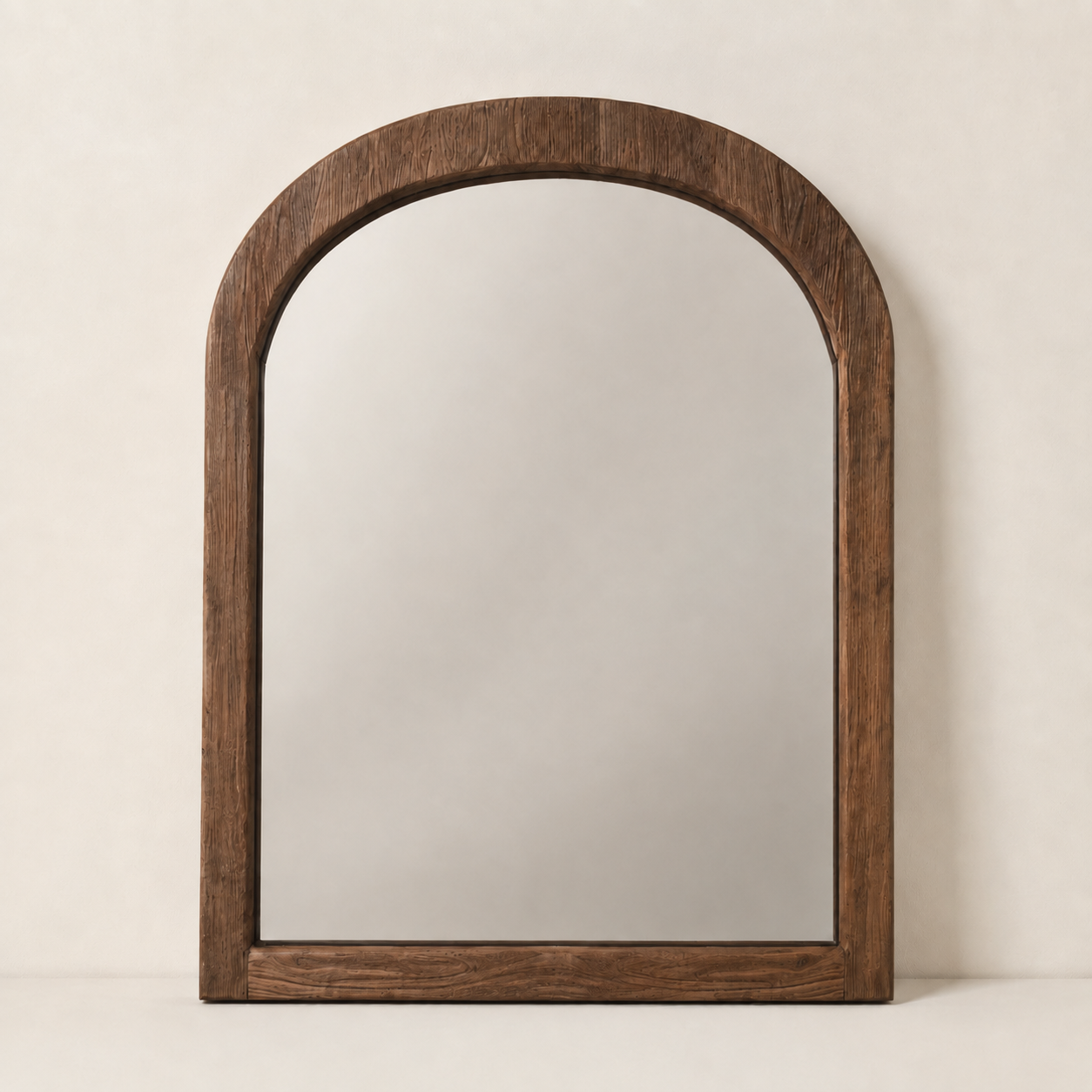 An arched wooden wall mirror with a thick, dark brown wood frame, standing against a neutral, warm off-white textured wall.