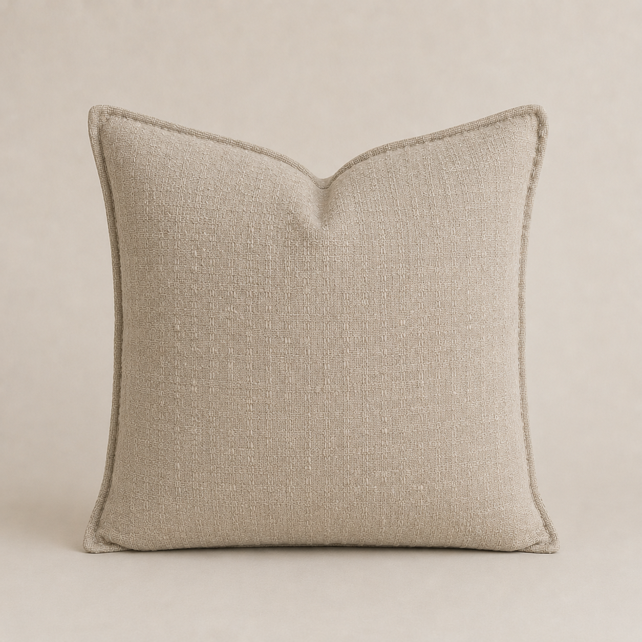 A square throw pillow in a textured beige linen fabric with a subtle V-shaped pleat at the top, finished with a matching beige piped edge, set against a matching neutral background.