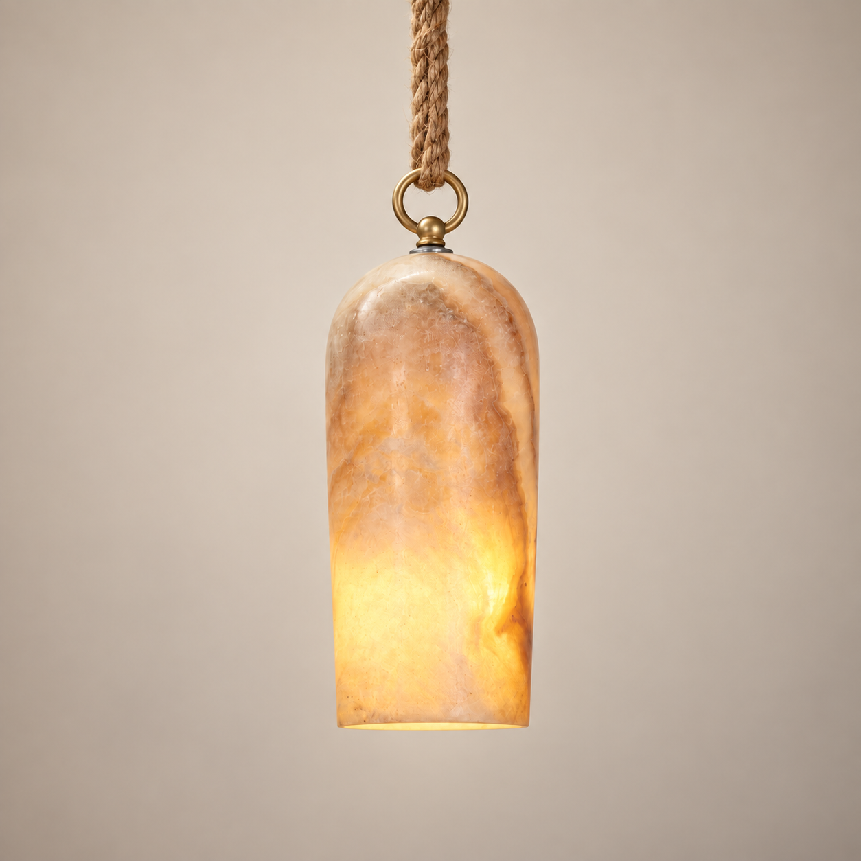 A modern pendant light with a cylindrical, rounded-top shade made of warm-toned marbled stone or resin, glowing with a soft warm light. It hangs from a thick braided rope cord with a brass ring and cap, set against a solid neutral beige background.