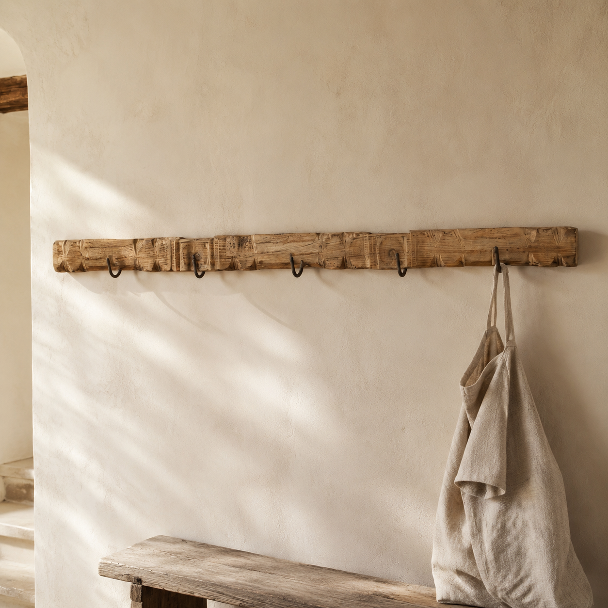 A rustic wooden wall-mounted coat rack with five black metal hooks, shown hanging a beige linen apron in a softly lit interior with a textured plaster wall and a wooden bench below.