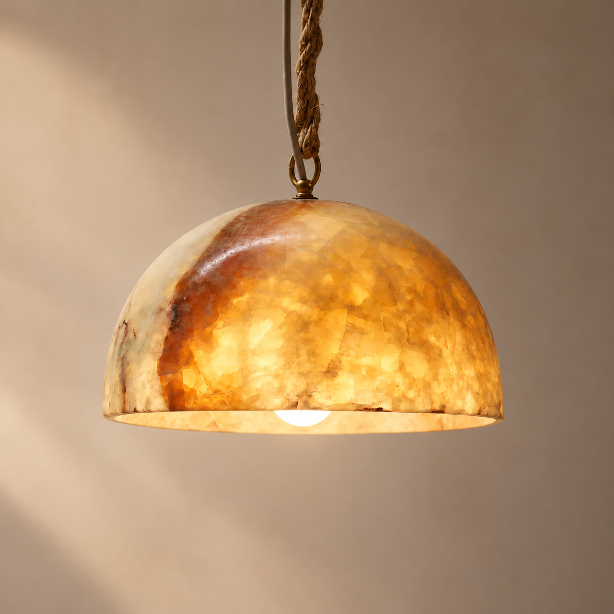 A dome-shaped pendant light shade made of warm-toned marbled stone or resin, glowing with a warm amber light. The shade hangs from a braided rope chain against a soft beige wall.