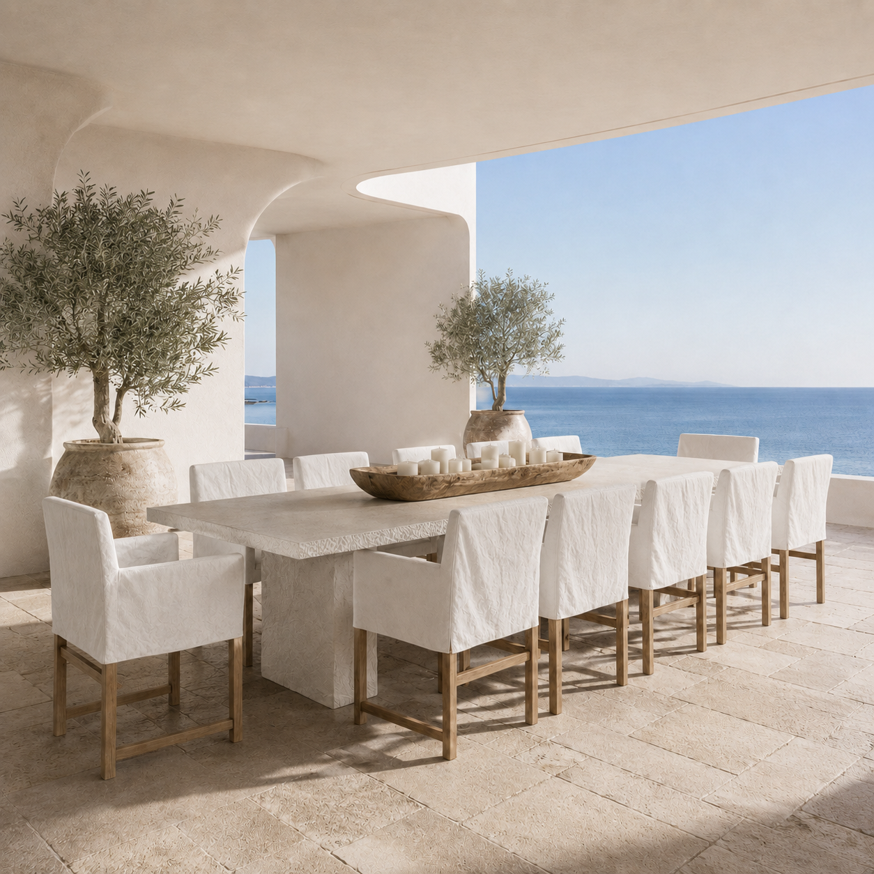 A minimalist outdoor dining set featuring a rectangular stone table and ten upholstered chairs with white fabric and light wood frames, arranged on a stone patio overlooking the blue sea under a covered terrace.