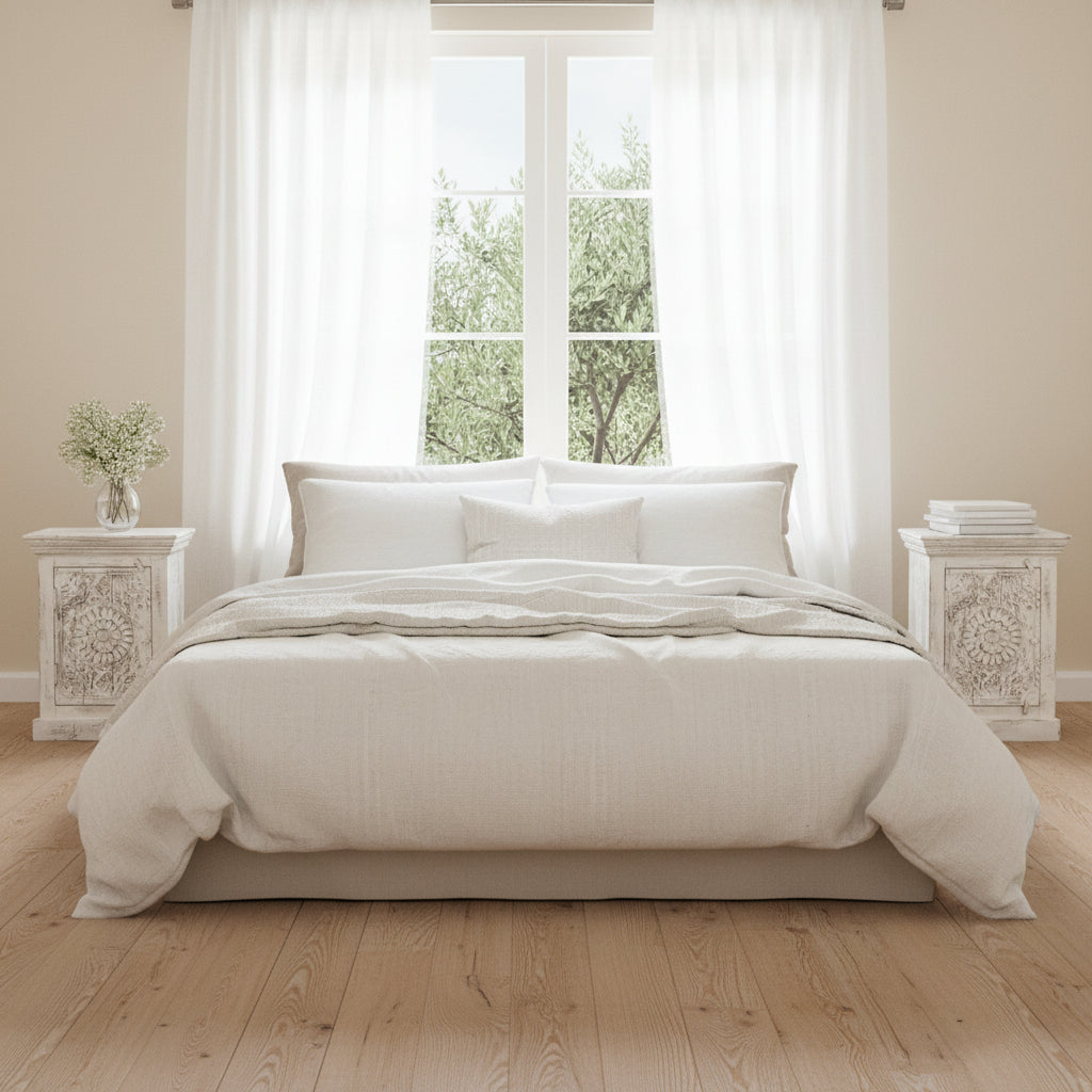 Neatly made bed with beige bedding in a bright bedroom with large windows.