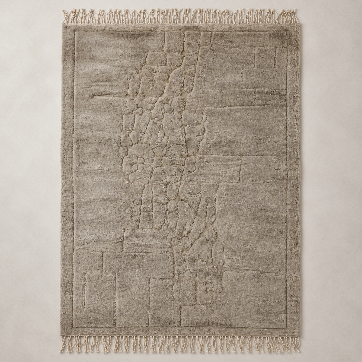 A rectangular area rug with a textured, abstract stone-like pattern in warm greige and beige tones, featuring fringed edges on all sides, shown flat against a neutral background.