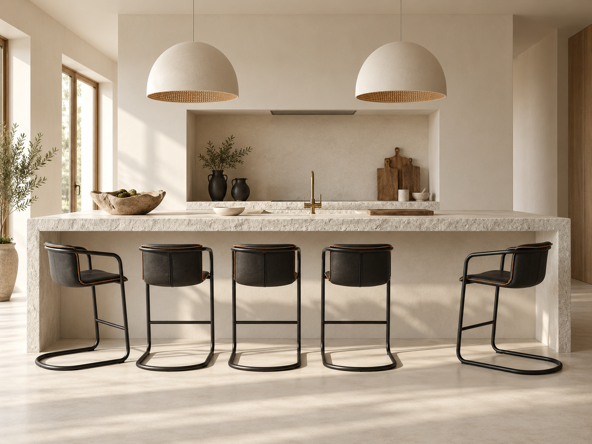 A modern kitchen island with a light stone countertop and four black bar stools, set in a bright, minimalist kitchen with pendant lights and warm natural light.
