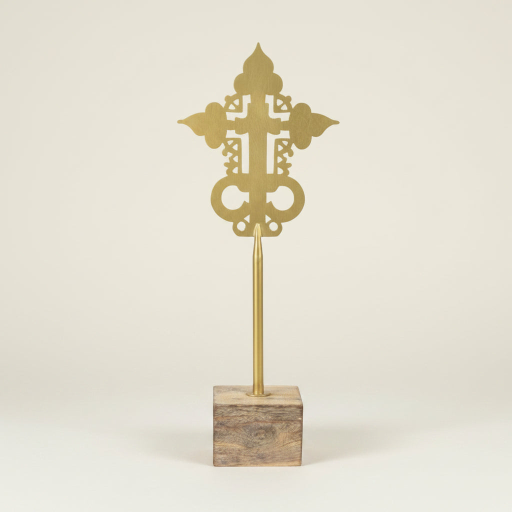 A decorative Orthodox cross finial in polished brass mounted on a square wooden base, photographed against a clean, light beige studio background.