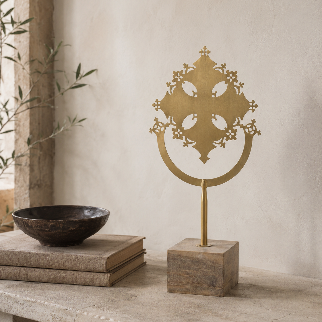 A decorative brass wall sculpture with an ornate filigree cross design on a circular base, mounted on a square wooden pedestal. The sculpture is styled on a stone ledge next to a dark ceramic bowl and books against a textured beige plaster wall.