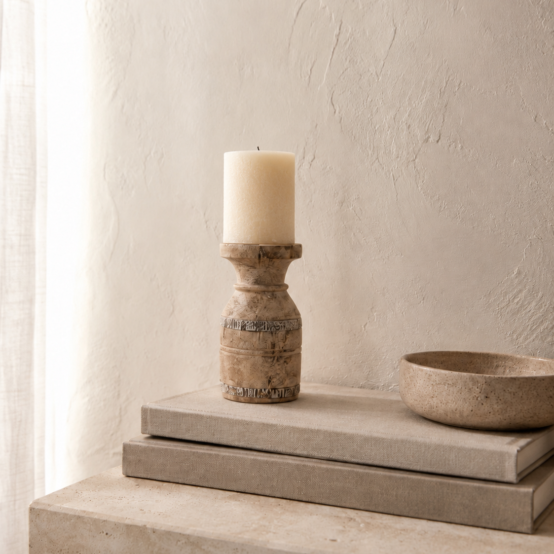 A rustic wooden candle holder with a cream pillar candle, styled on stacked stone slabs next to a stone bowl against a textured beige plaster wall.