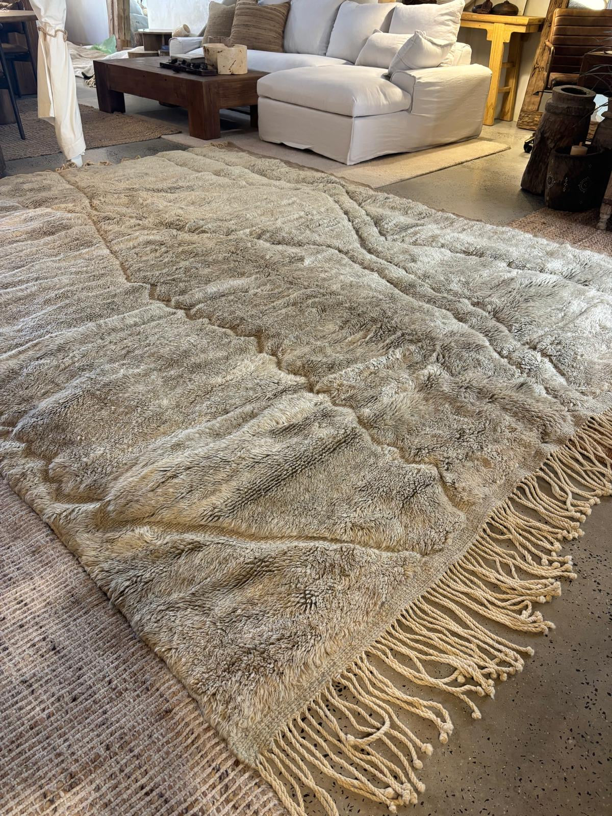 A hand-selected Moroccan Berber rug with a beige and greenish hue and fringes, laid out on the floor in a room.
