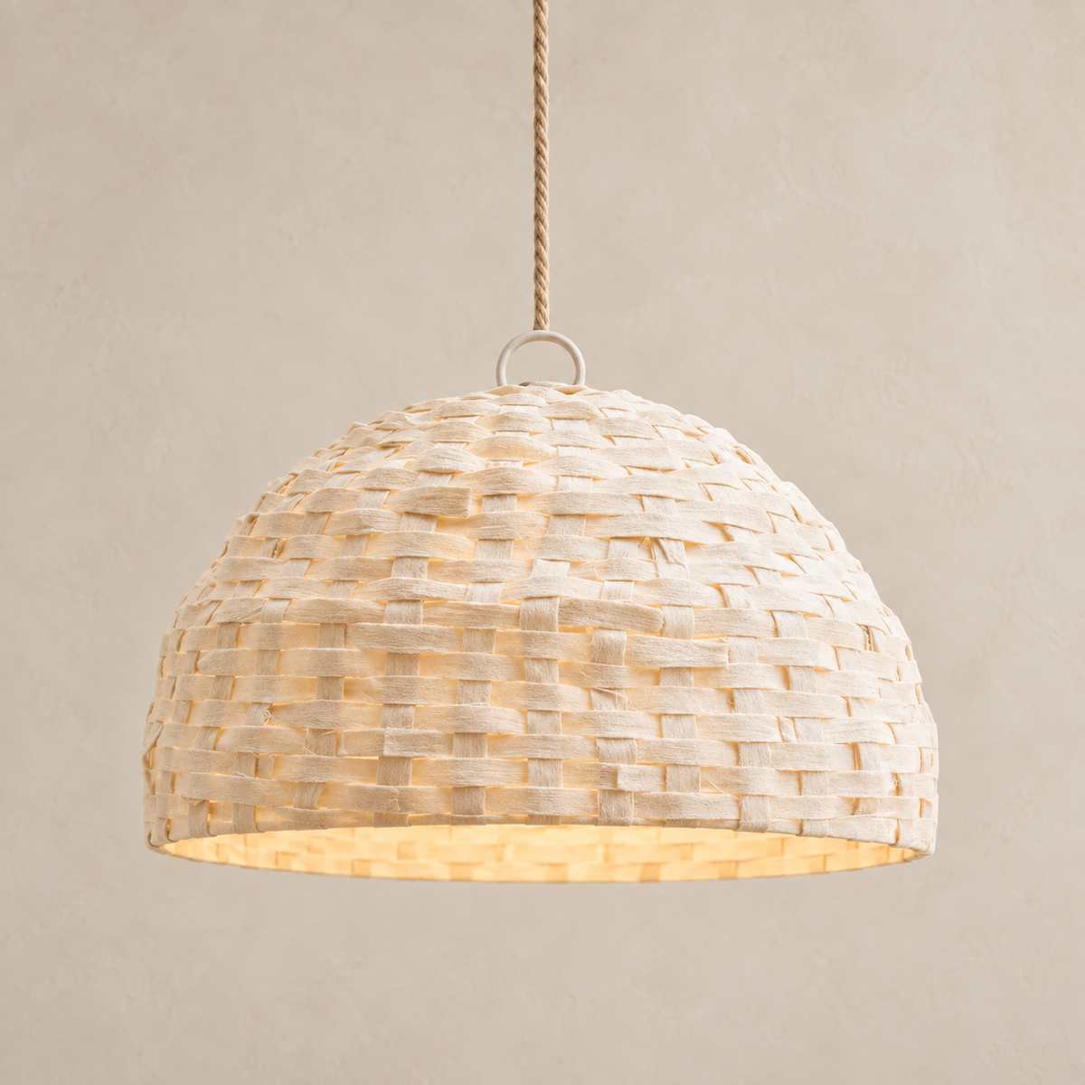 A dome-shaped woven pendant light made from natural beige raffia or straw strips, suspended by a rope cord against a warm beige textured wall.