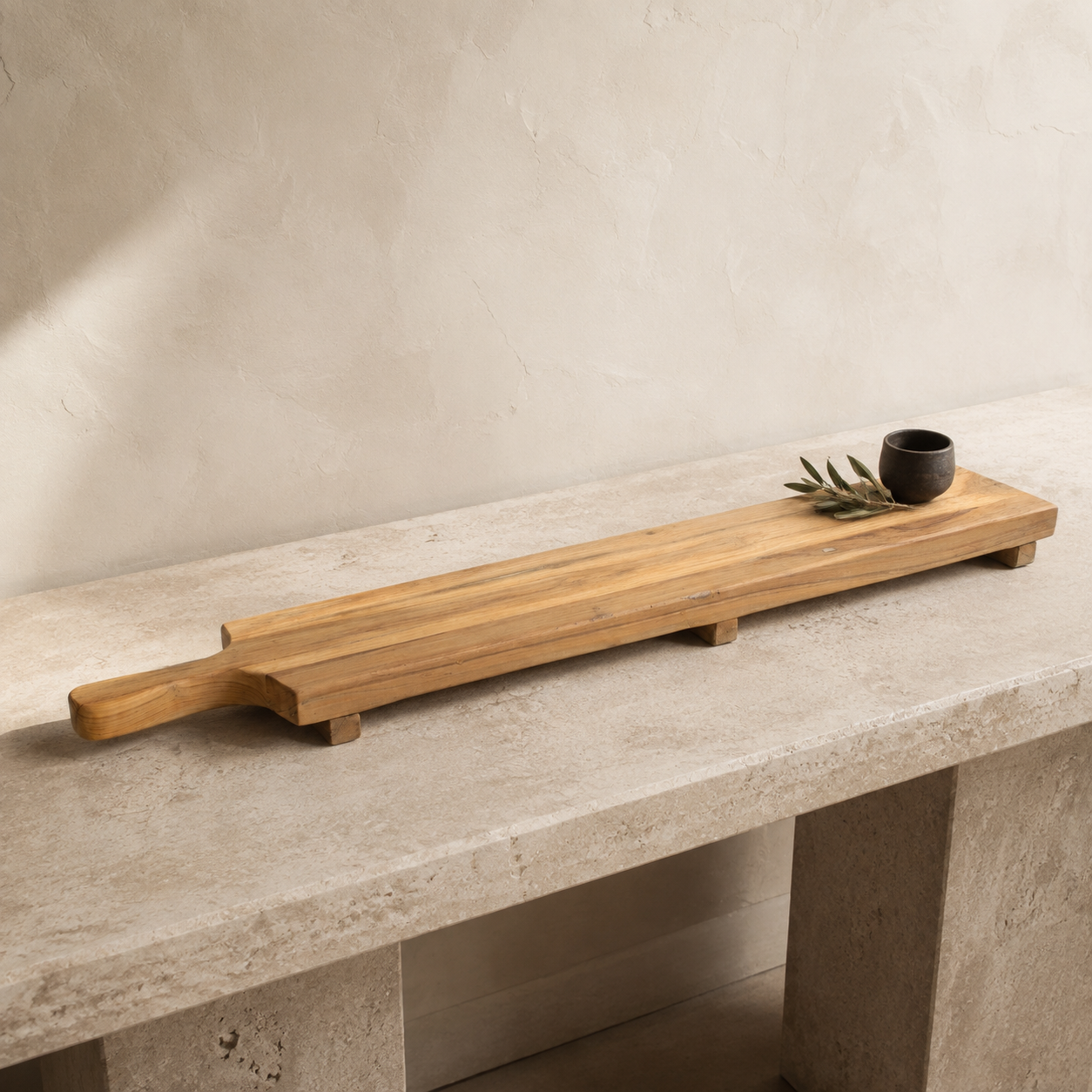 A long rectangular wooden serving board with a handle, placed on a stone table against a textured beige wall. A small dark cup and dried greenery rest on one end.