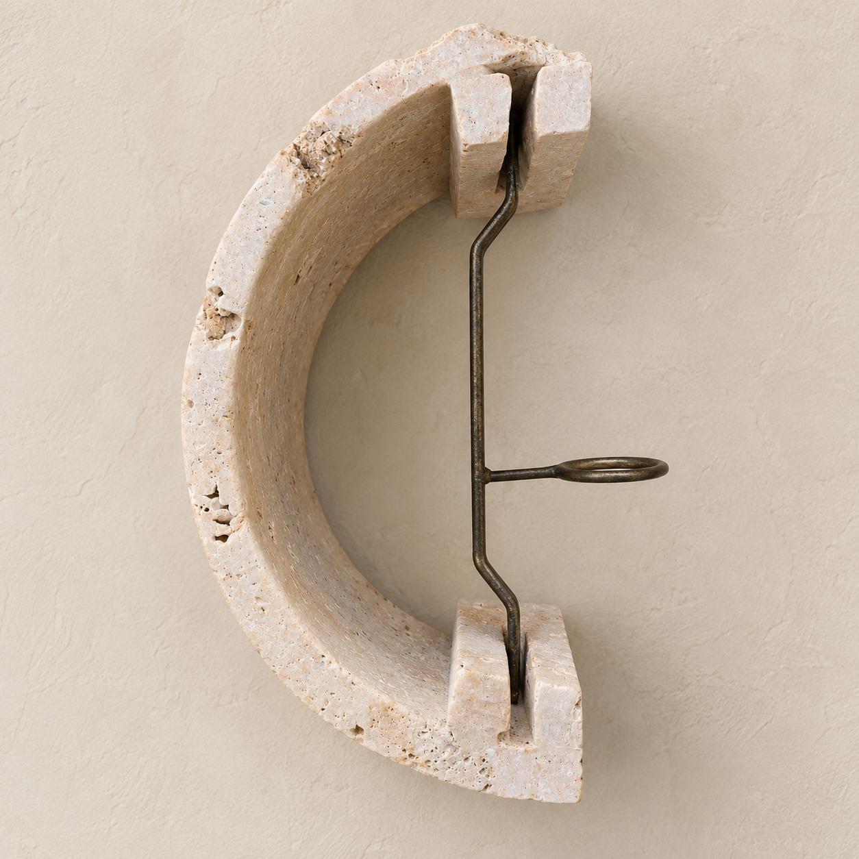 A modern wall-mounted crescent-shaped hook made of textured beige travertine stone with a dark metal tension wire element, shown against a matching textured beige plaster wall.