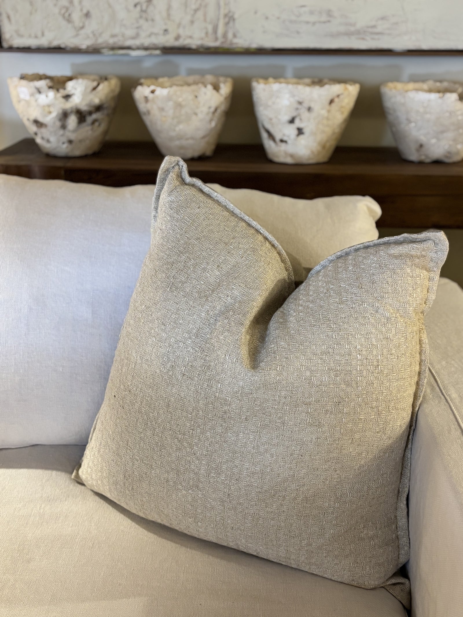 A beige cushion with a textured fabric, featuring hand stitching details, placed on a sofa with a shelf in the background containing decorative items.