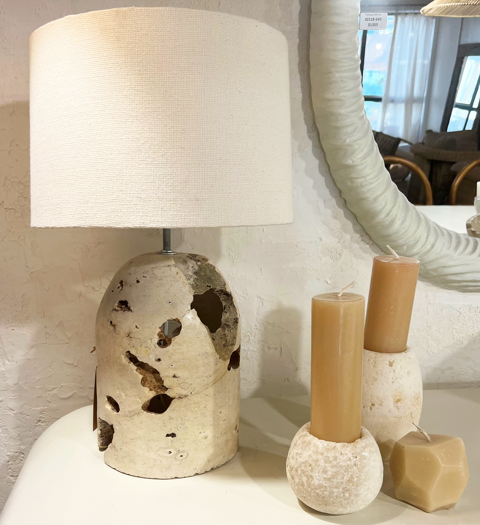 A table lamp with a textured cream fabric drum shade and a porous beige stone base sits on a white surface next to two tan pillar candles in matching stone holders.
