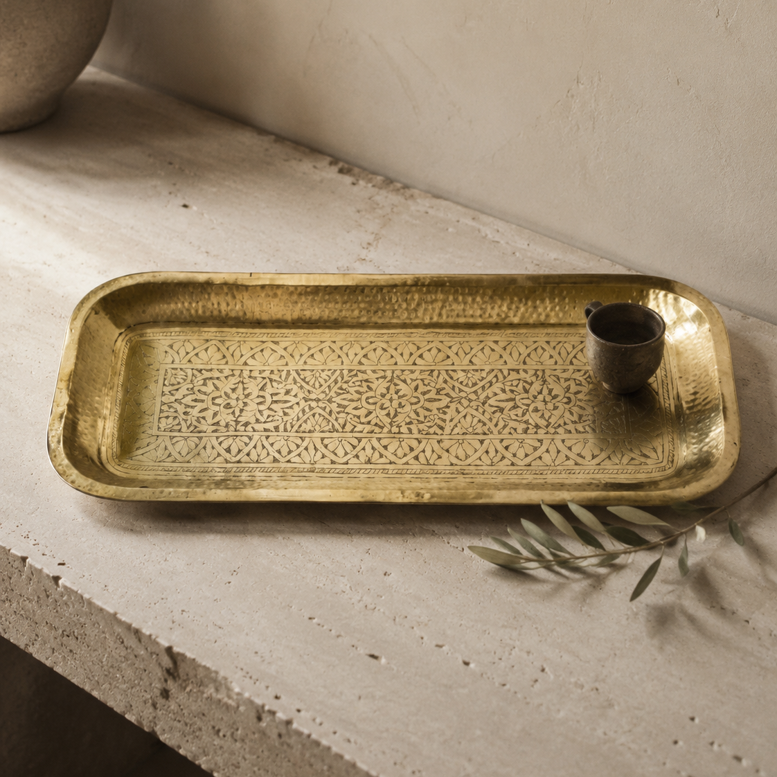 A rectangular hammered brass tray with intricate cutout patterns, resting on a textured concrete ledge next to a small dark metal cup and a sprig of olive leaves.