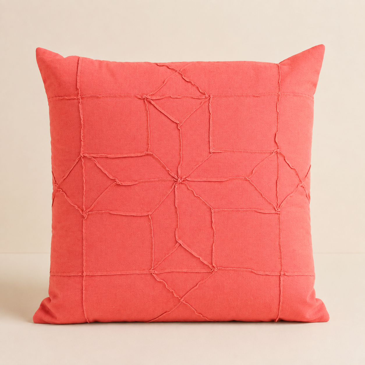A square coral pink throw pillow with a geometric, stitched quilted pattern on the front
