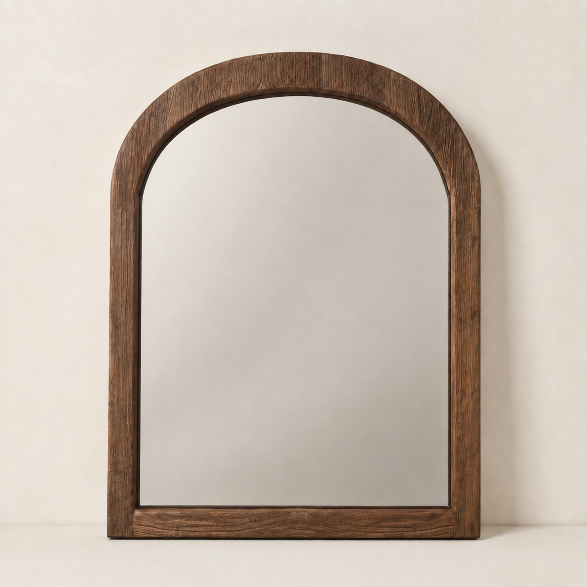 An arched wooden wall mirror with a thick, dark brown wood frame, standing against a neutral, warm off-white textured wall.