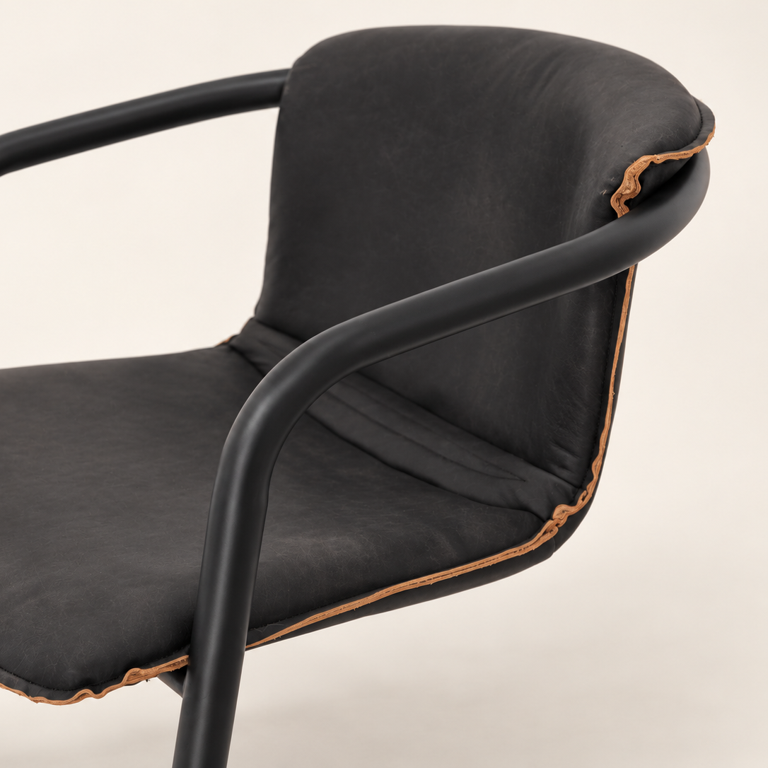A modern accent chair with a black upholstered seat and backrest, supported by a matte black metal frame with curved armrests, featuring contrasting tan leather raw edge.