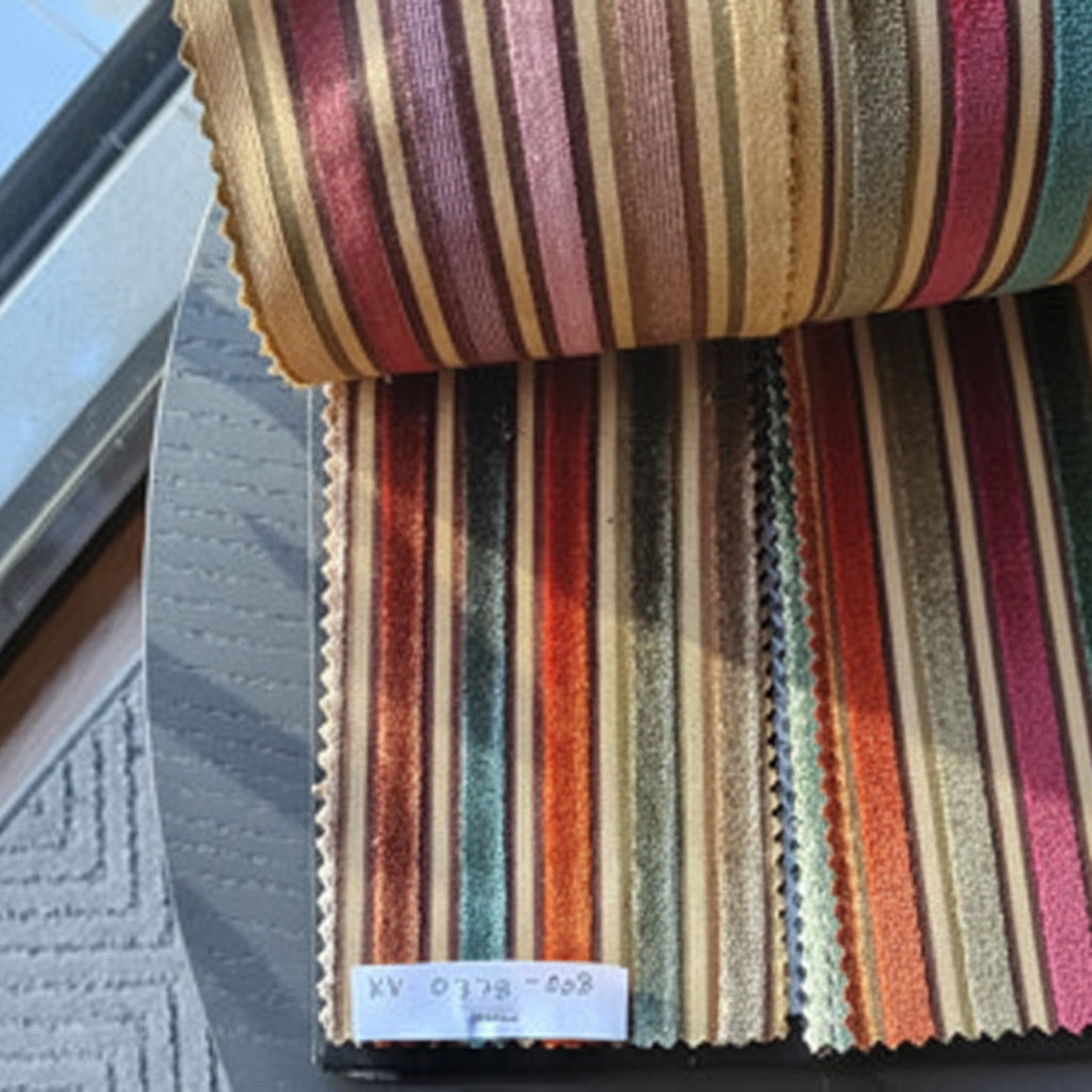 A close-up of striped fabric swatches in multiple colors, including gold, red, orange, green, blue, and brown, with a small white label at the bottom.