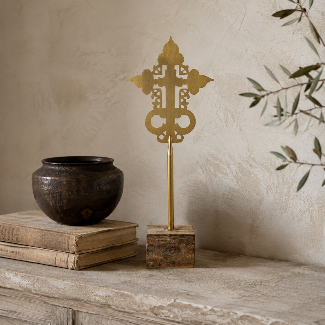 A decorative brass cross finial on a slender stand with a square wooden base, displayed on a stone ledge beside stacked books and a dark ceramic pot against a textured beige wall.
