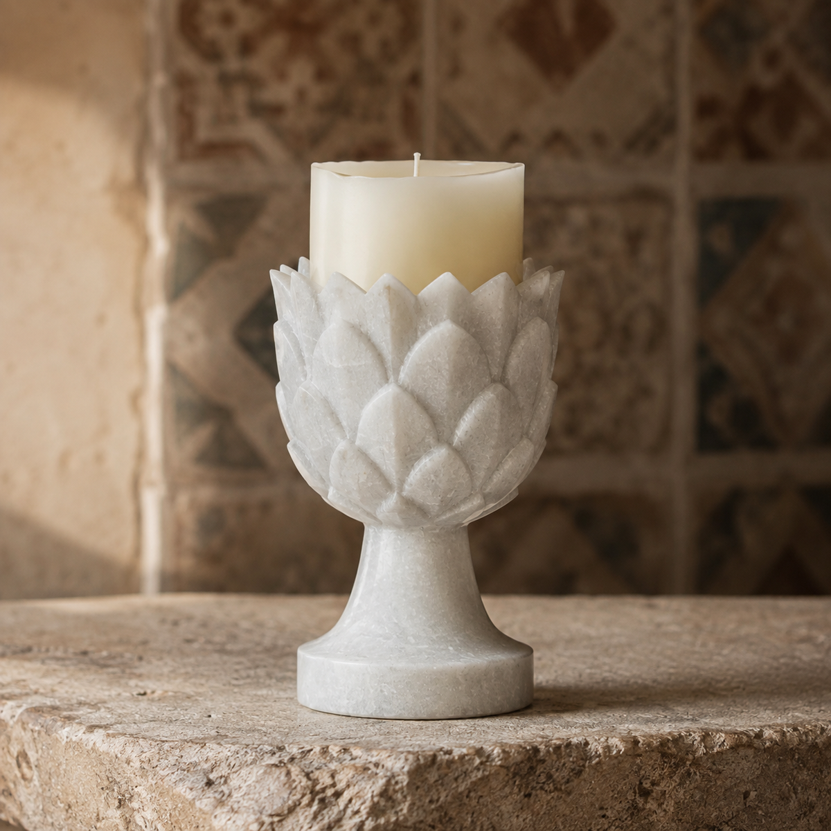 A white textured stone candle holder with a lotus petal design, holding a cream-colored pillar candle, set on a rustic stone surface with a blurred tiled background.