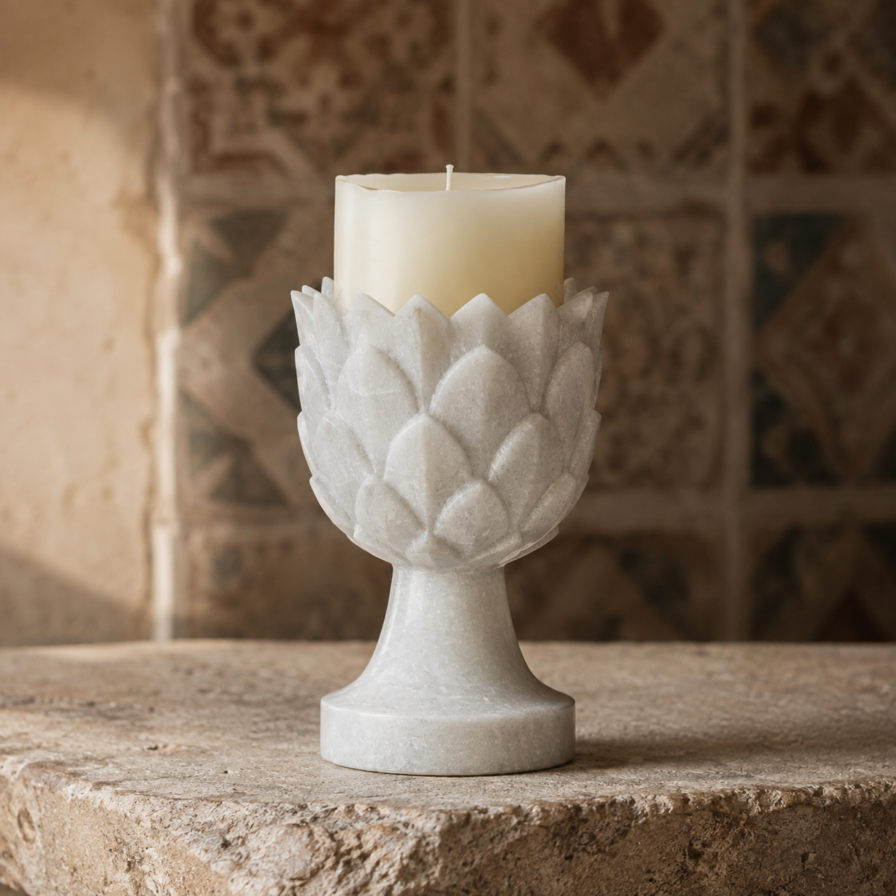 A white textured stone candle holder with a lotus petal design, holding a cream-colored pillar candle, set on a rustic stone surface with a blurred tiled background.