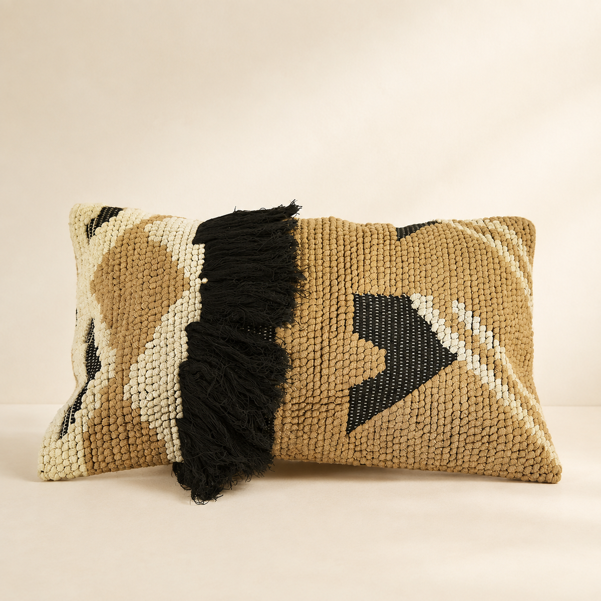 A rectangular woven throw pillow in a tan and cream geometric pattern with a prominent black fringe detail, set against a warm beige background.