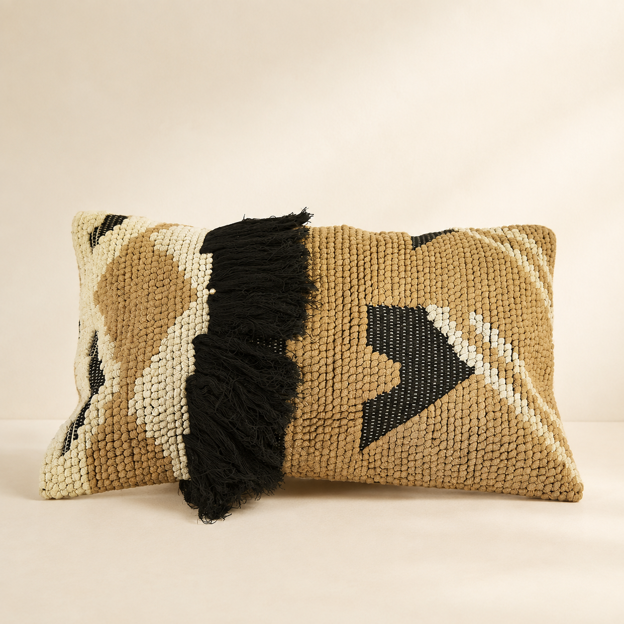A rectangular woven throw pillow in a tan and cream geometric pattern with a prominent black fringe detail, set against a warm beige background.