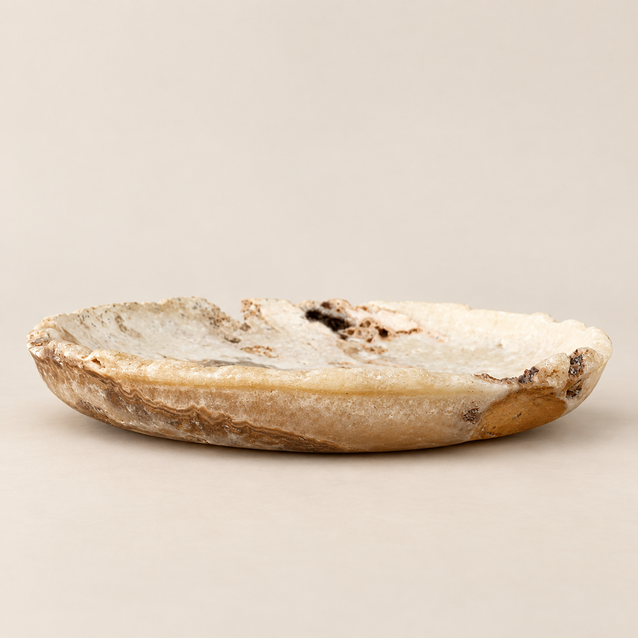 A small, shallow dish carved from natural agate or quartz with a rough, crystalline texture and organic brown and black inclusions, shown on a neutral beige background.