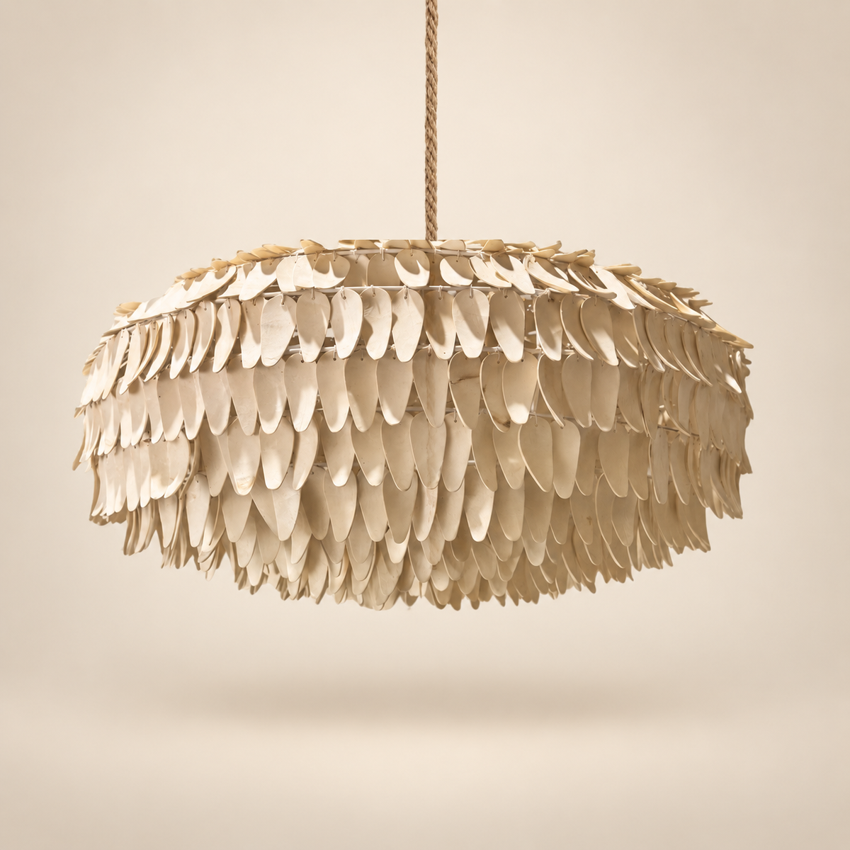 A modern pendant light with a round shade made of overlapping cream-colored petal or shell-like pieces, suspended by a twisted rope cord against a warm beige background.