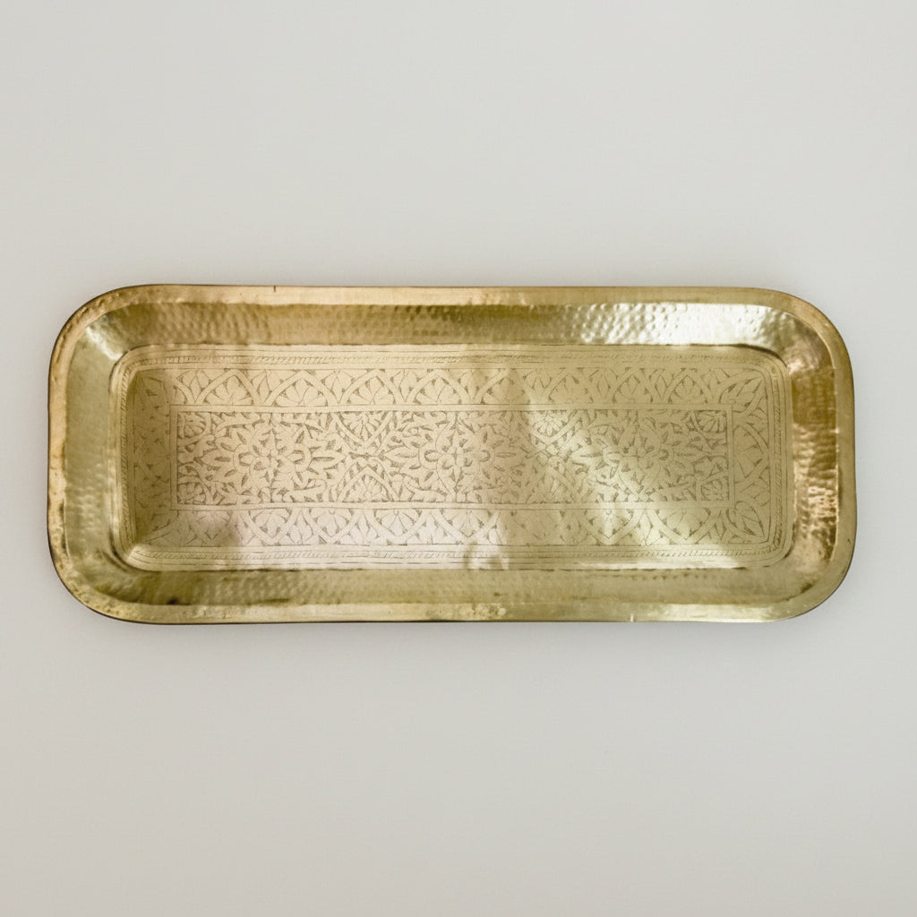 A rectangular brass tray with rounded corners and an embossed floral pattern, displayed on a textured off-white plaster background.