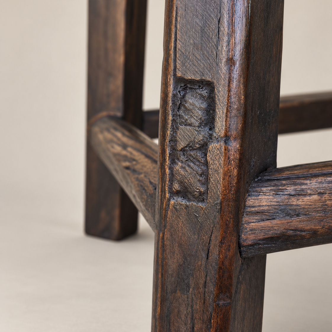 Close-up of a dark brown wooden stool leg showing a carved rectangular recess and detailed wood grain texture against a neutral beige background.