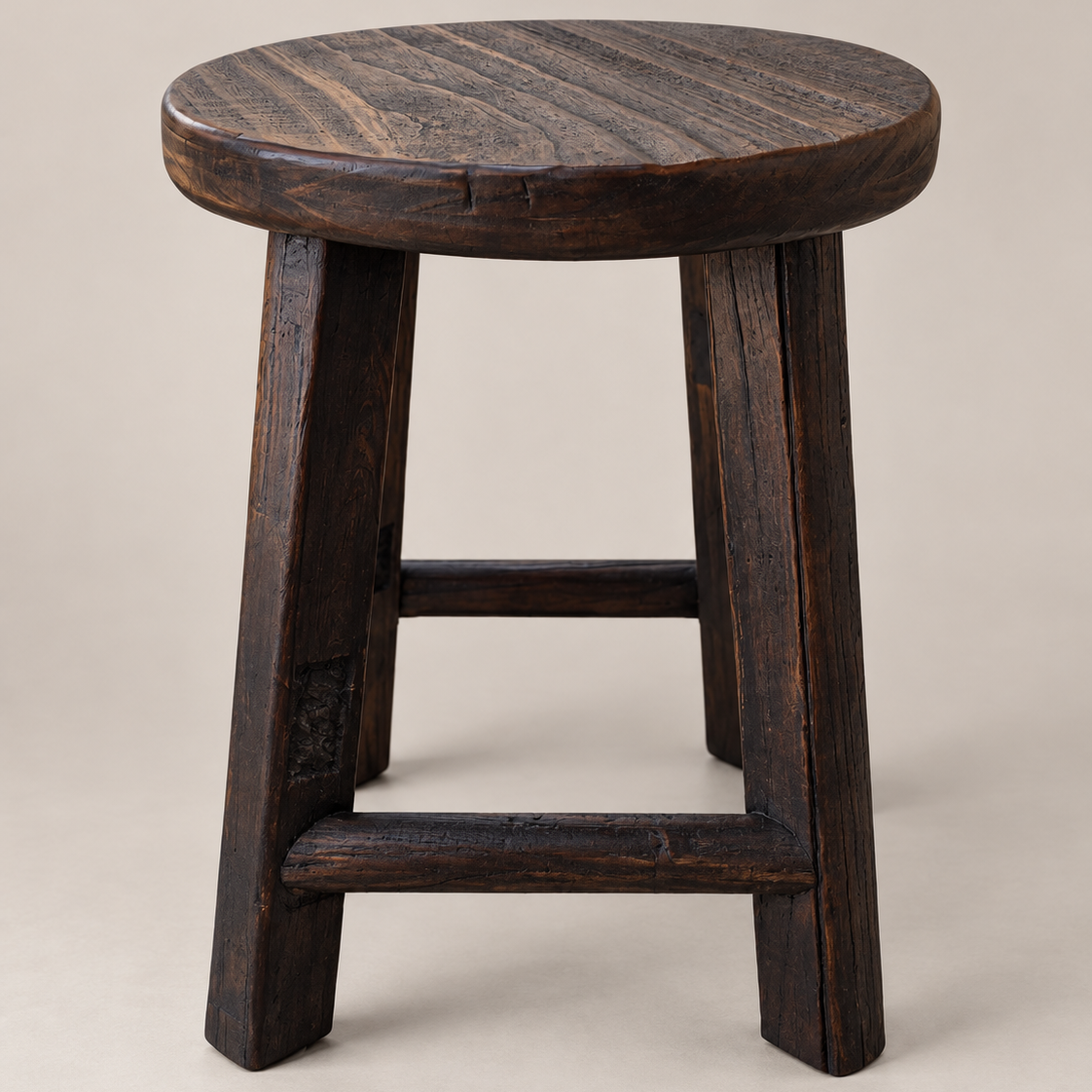 A rustic dark brown wooden stool with a round seat, four sturdy legs, and a front support bar, photographed against a plain light beige studio background.