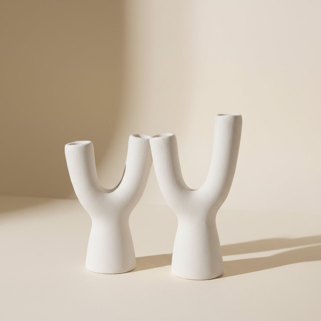 A white ceramic Y-shaped vase or sculptural holder displayed among neutral-toned decorative objects, including a small wooden bowl and dried botanical elements.