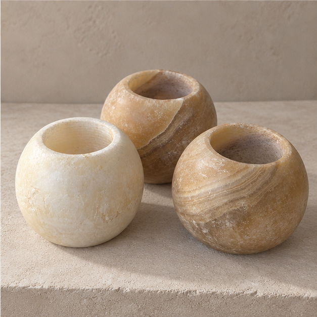 Three small round stone vessels with open tops, shown on a textured beige surface against a matching neutral background. Two vessels have warm tan and brown marbled stone, and one has a creamy off-white marbled stone.