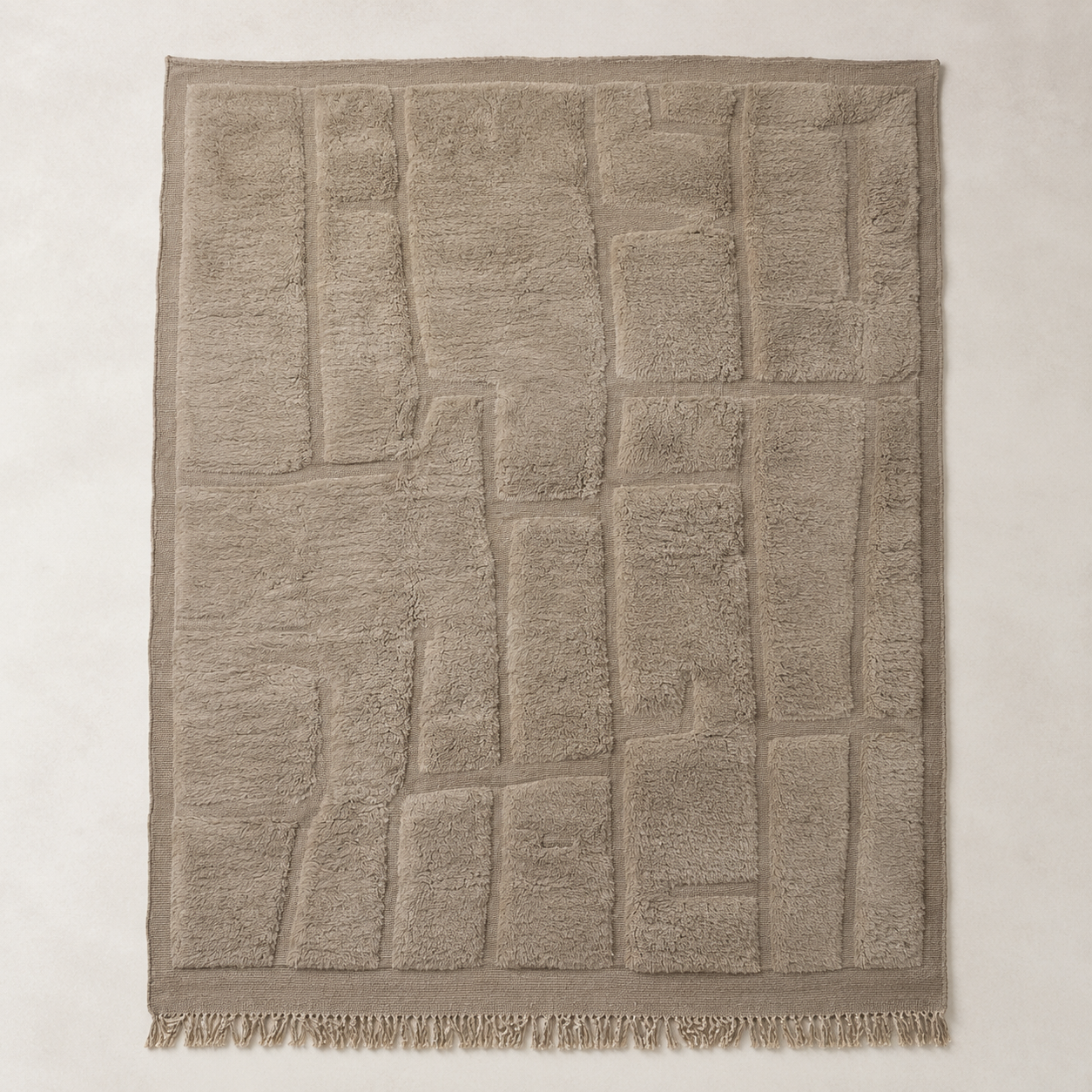 A rectangular beige textured area rug with a raised geometric block pattern and fringed edges, shown flat against a neutral light beige background.