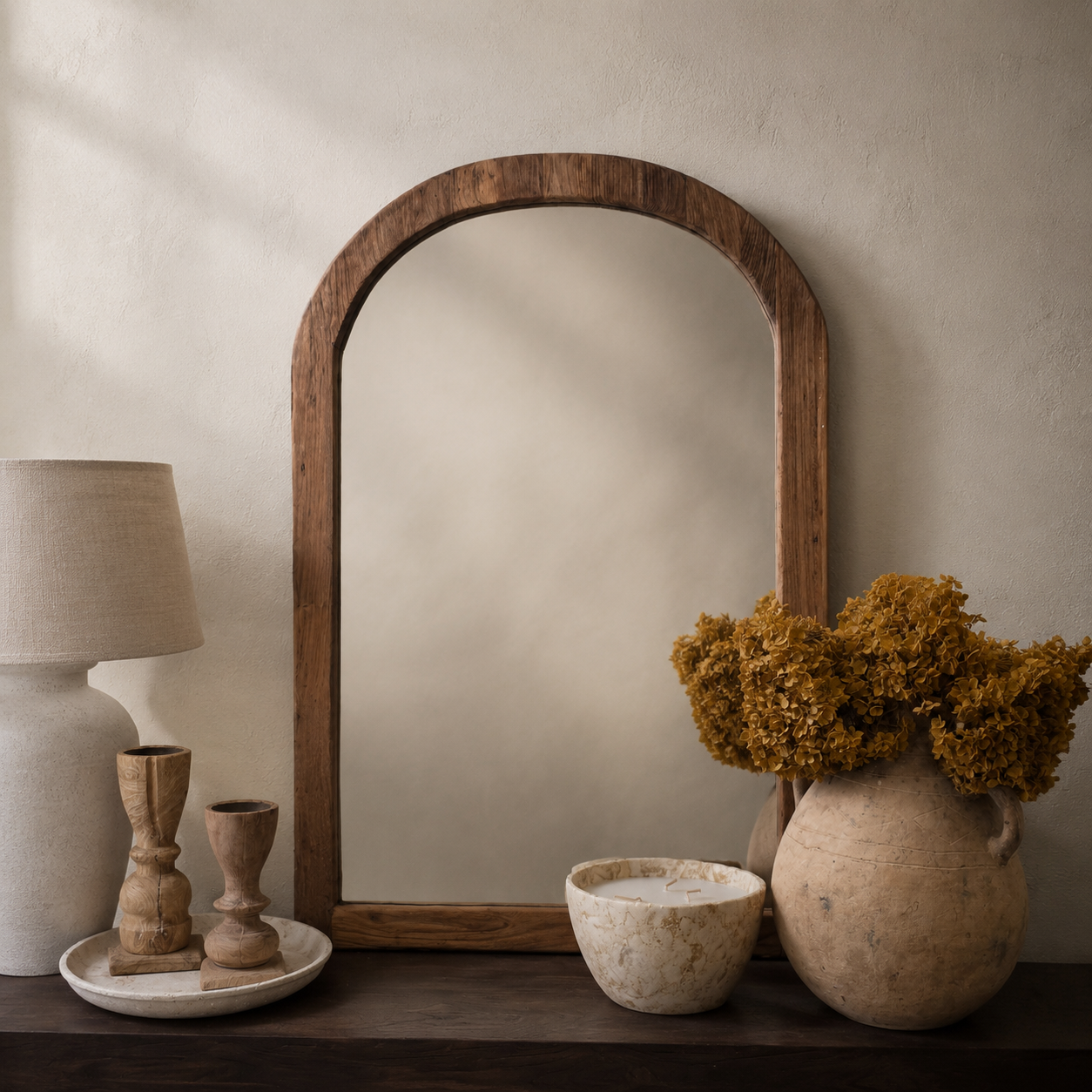 A large arched wooden mirror with a rustic dark brown frame, styled on a dark wooden shelf next to a table lamp, small wooden candle holders, a ceramic bowl, and a large vase with dried yellow flowers against a textured beige wall.