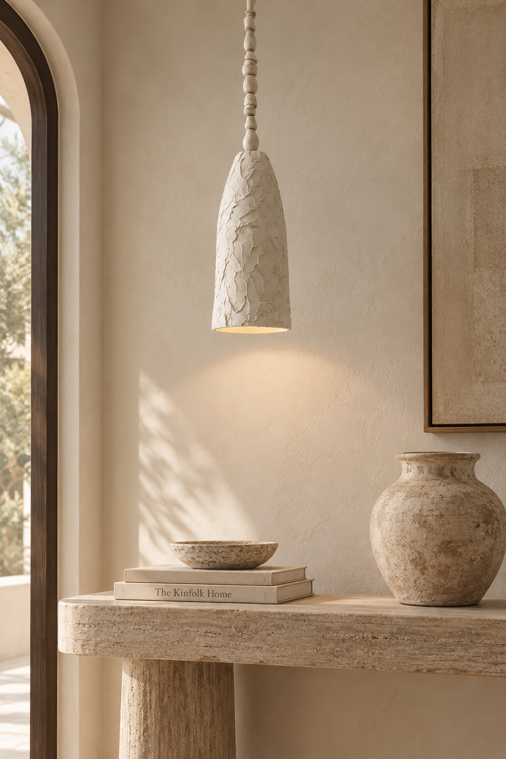 A textured stone pendant light with a conical shade hangs over a rustic stone console table in a warm, neutral-toned interior. The scene includes a ceramic vase, stacked books, and a framed artwork on a beige plaster wall.