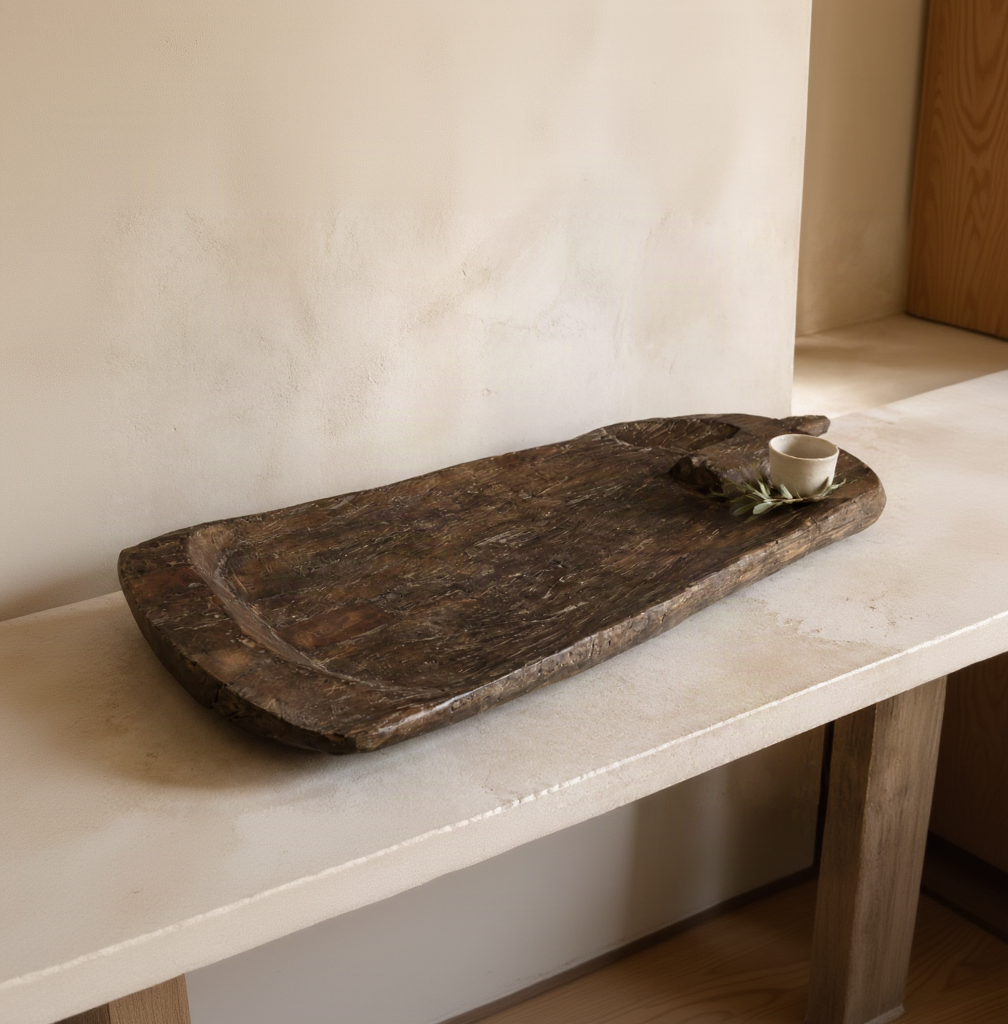 A rustic dark brown wooden serving tray with a carved bowl, displayed on a light stone table against a textured beige plaster wall, with a small white cup and green leaves as props.