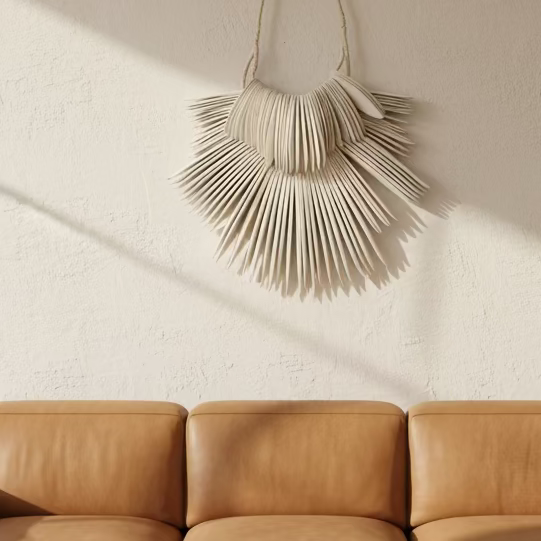 A tan leather three-seat sofa in a modern living room setting with a textured cream wall, a woven rug, and a hanging woven wall decor piece above.